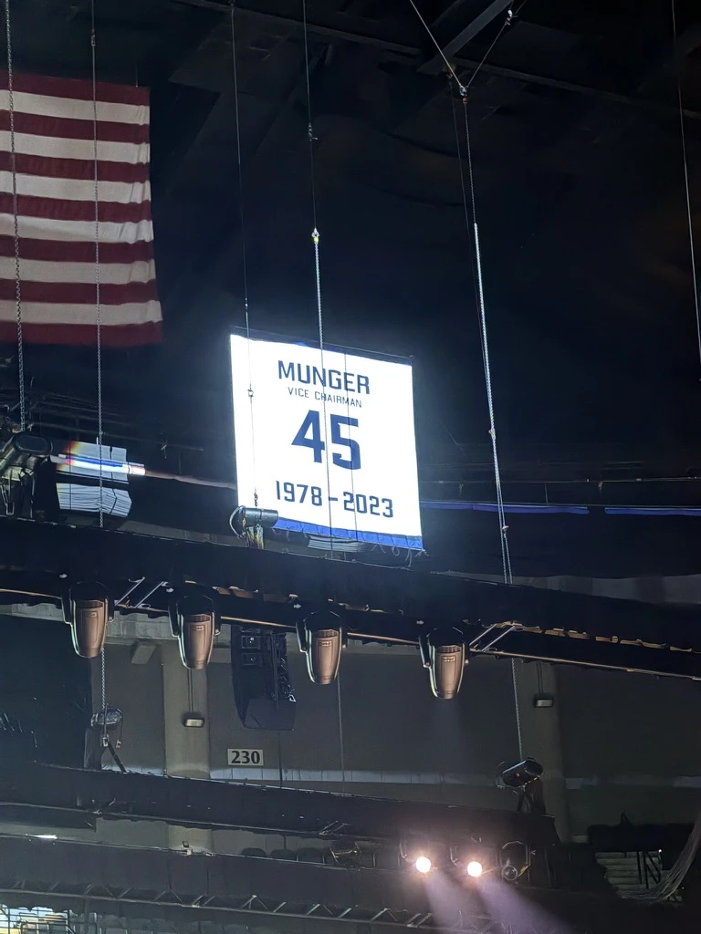 A white Charlie Munger tribute banner hangs high inside the arena beside an American flag.