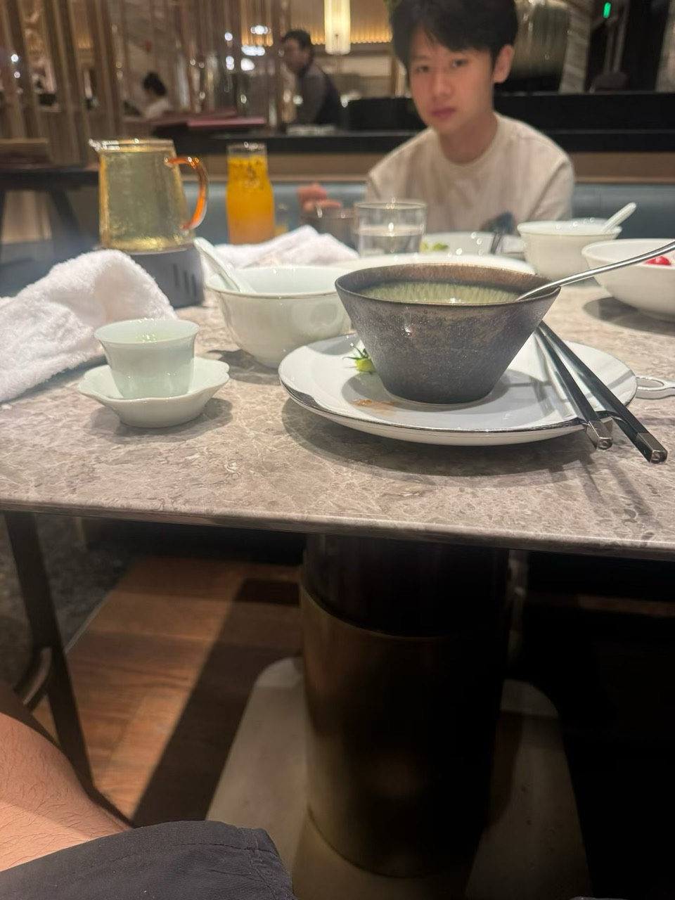 A bowl of soup, tea, and tableware in the foreground, with a friend seated softly out of focus across the restaurant table.