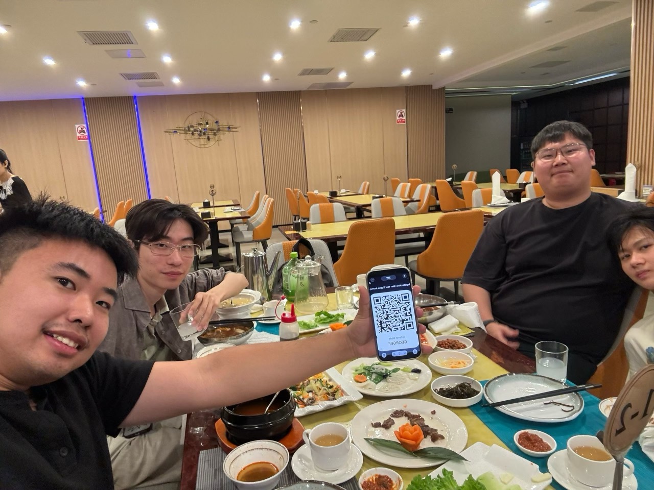 Four friends gather around a restaurant table covered with dishes and tea, leaning into a group photograph after dinner.