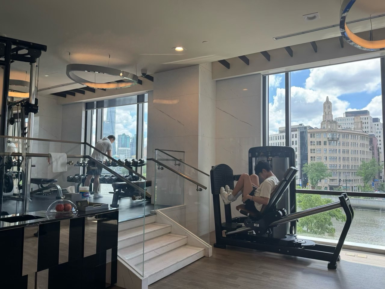 A friend uses a leg-press machine in a bright gym overlooking the river and city buildings through large windows.