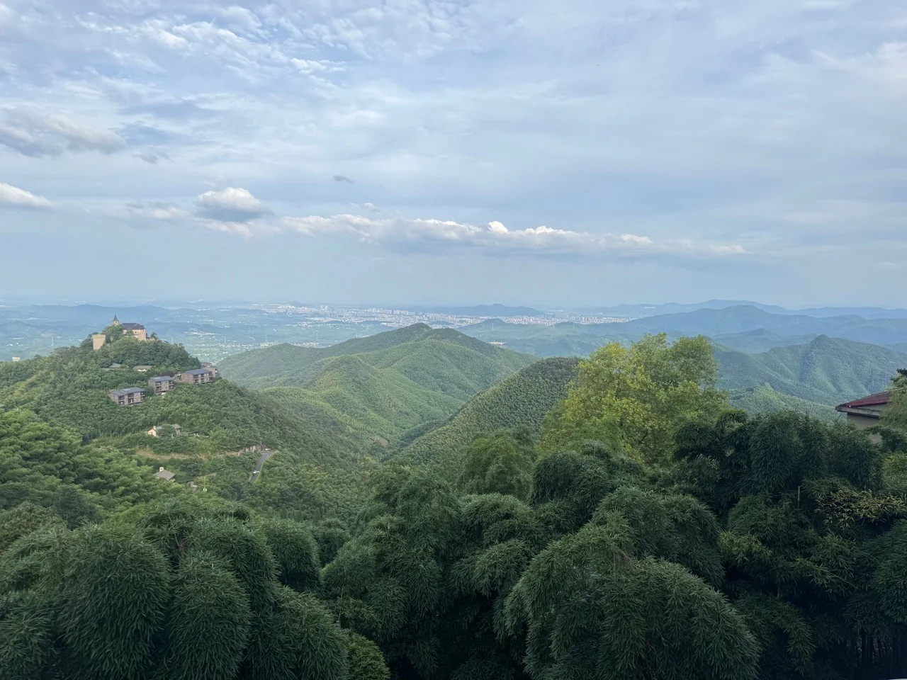 A wide daytime view across layered green ridges and bamboo forest in Moganshan beneath a pale blue sky.