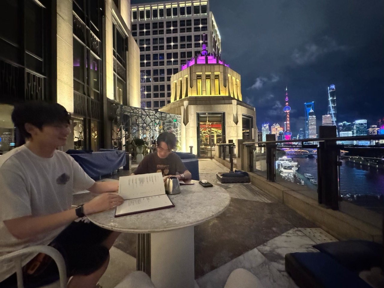 Two friends sit at a rooftop table at night, with the illuminated Shanghai skyline and river spread out behind them.